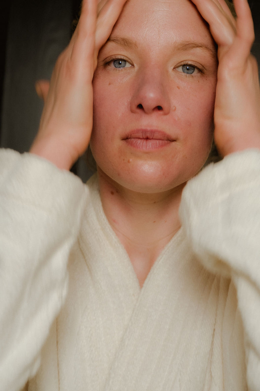 Person wearing a white robe with hands on head against a dark background